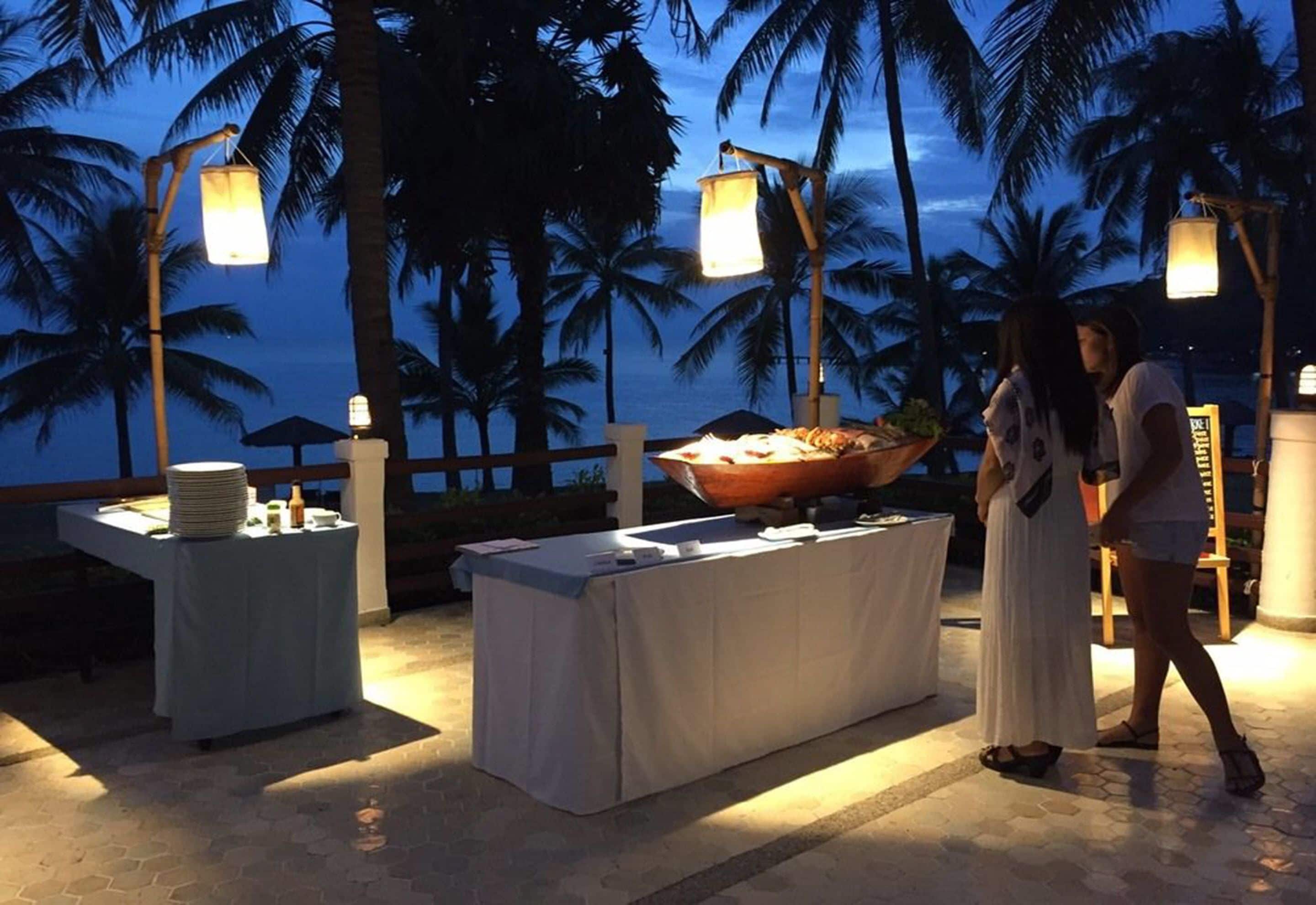 Un área de comedor al aire libre con mesas y sillas, una mesa de buffet y palmeras al fondo.