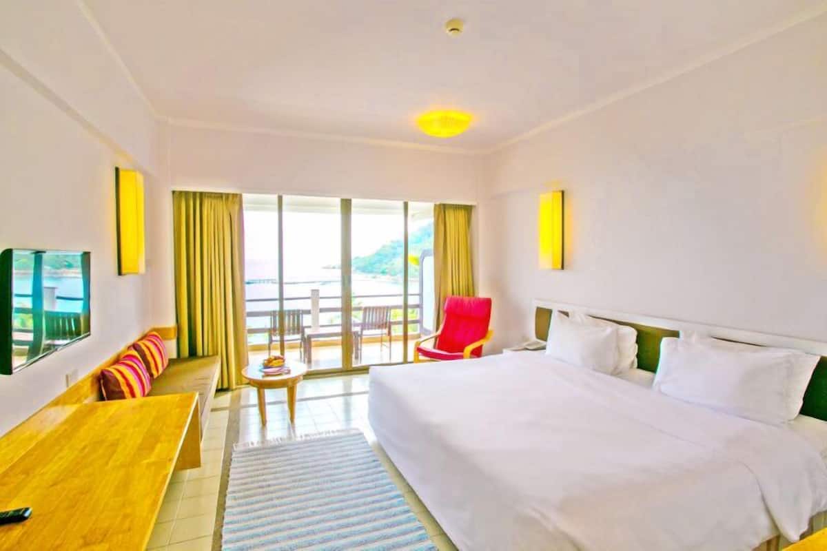 Habitación de hotel con una cama grande, un banco de madera, una mesita y una silla. Cuenta con un balcón con vistas al exterior.