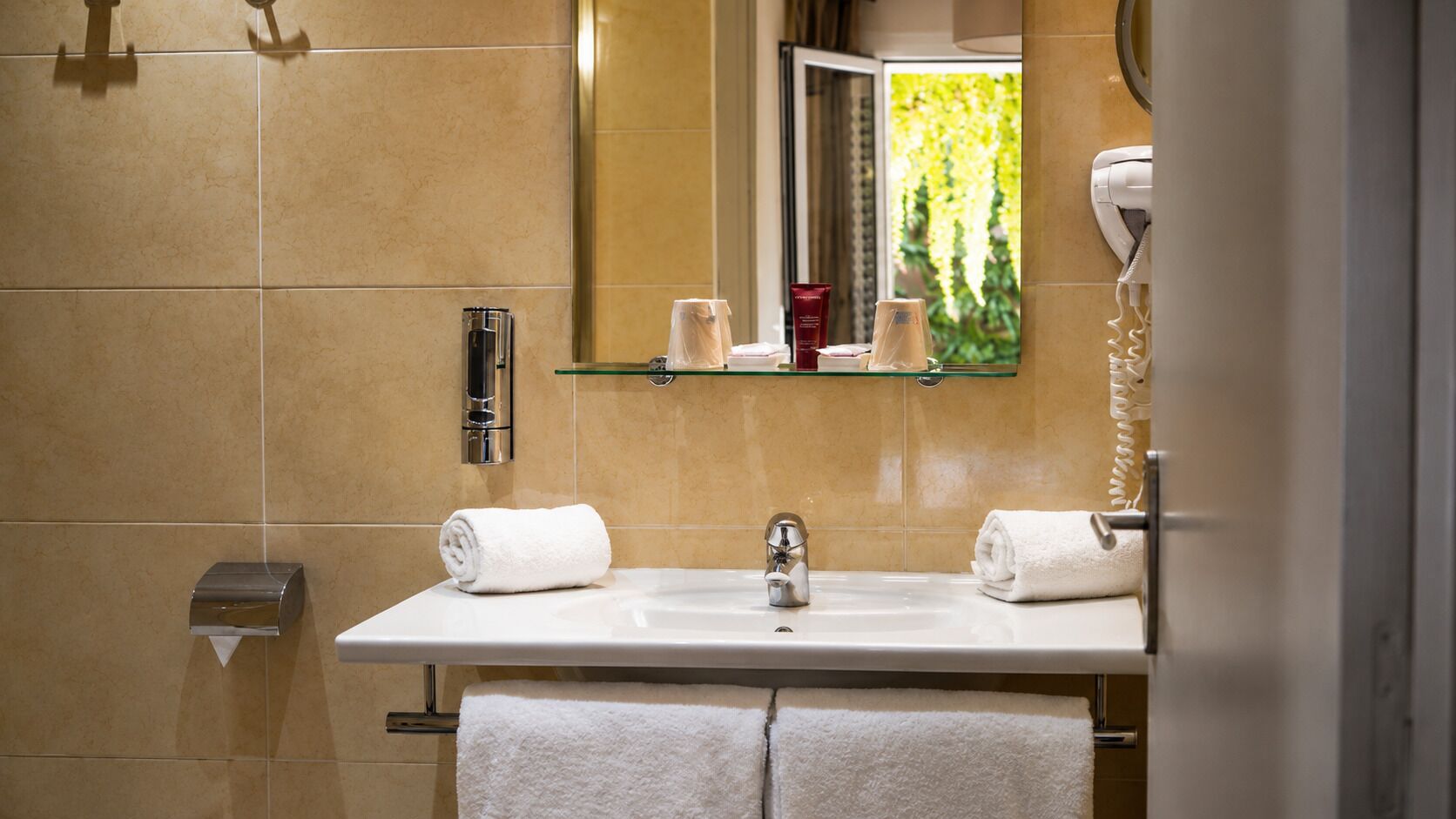 Een badkamer met een wit wastafel, twee handdoeken, een glazen plankje met toiletartikelen en een spiegel die groen reflecteert buiten.