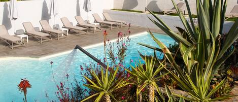 Un área de piscina con sillones de descanso y sombrillas, rodeada de plantas y un muro de piedra.