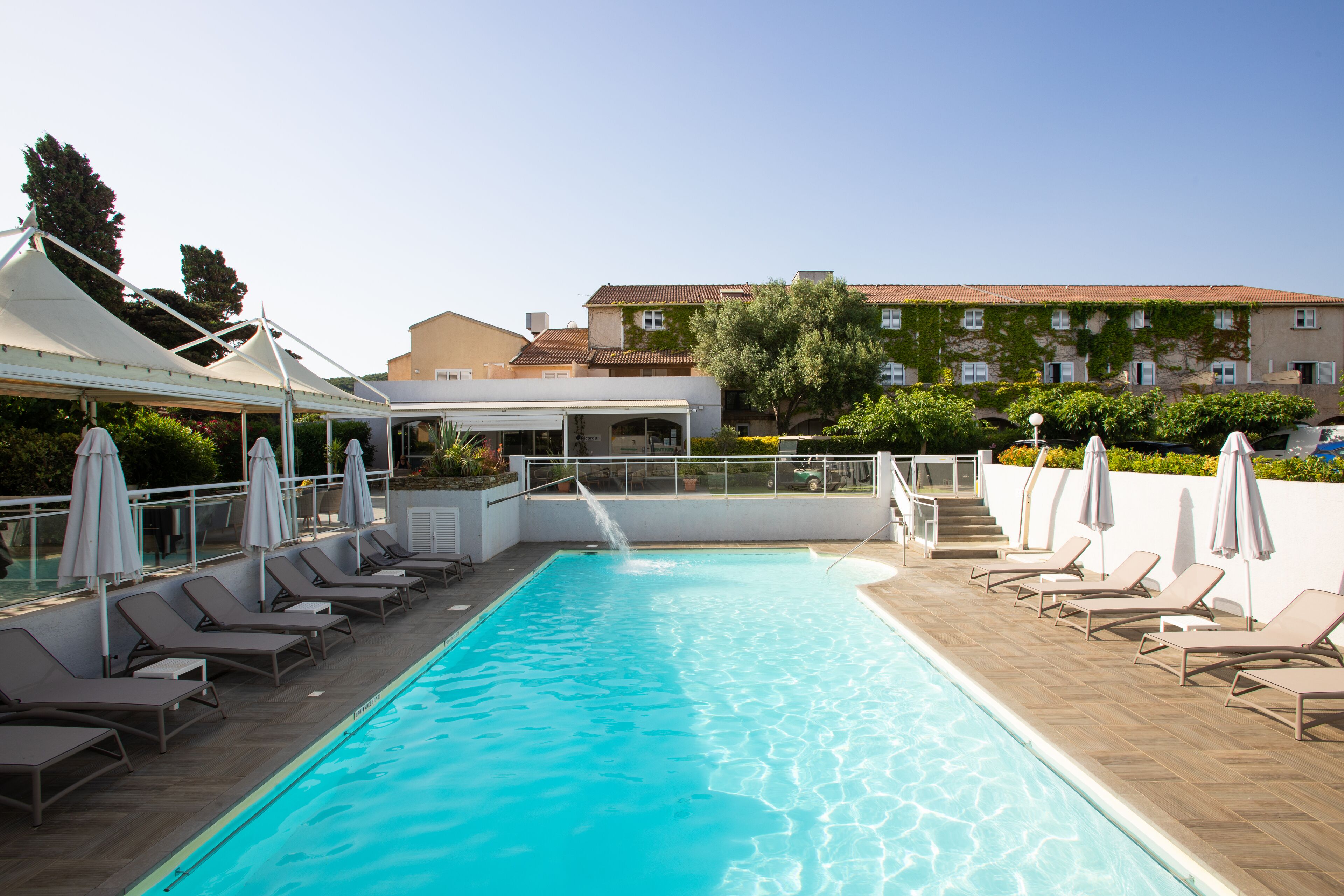 A swimming pool with lounge chairs and umbrellas, a covered area, and a building with a tiled roof in the background.