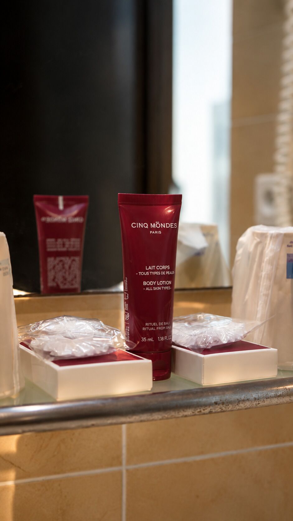 Una encimera de baño con una botella roja de crema corporal con la etiqueta 'Cinq Mondes Paris' y una caja blanca con una bolsa de plástico.