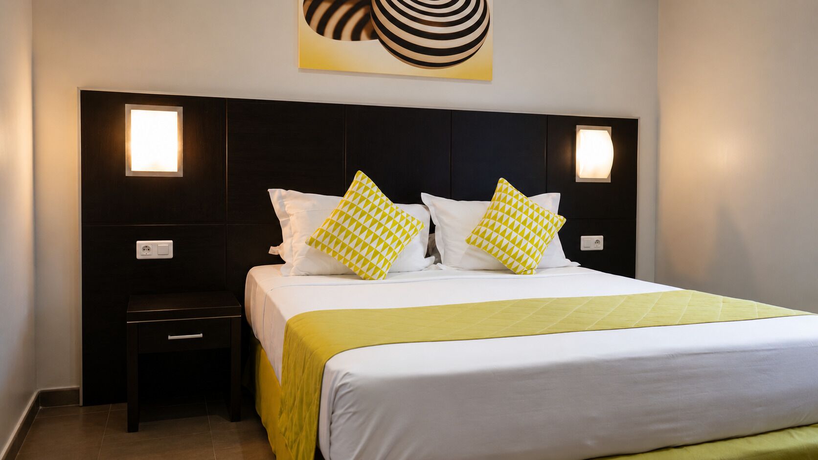 Habitación de hotel con cama, dos mesitas de noche, lámpara de pared y una obra de arte en la pared.