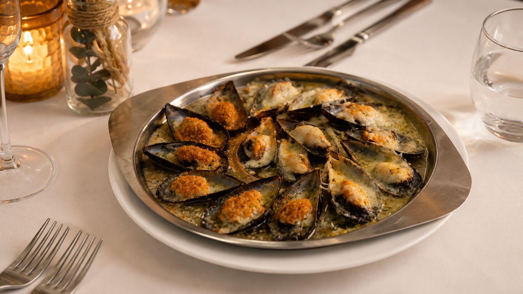 A plate of mussels with breadcrumbs on a table with a candle and a glass of water.
