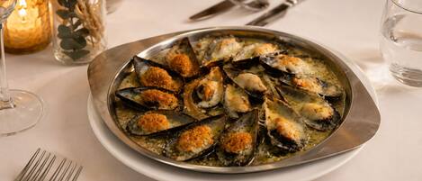A plate of mussels with breadcrumbs on a table with a candle and a glass of water.