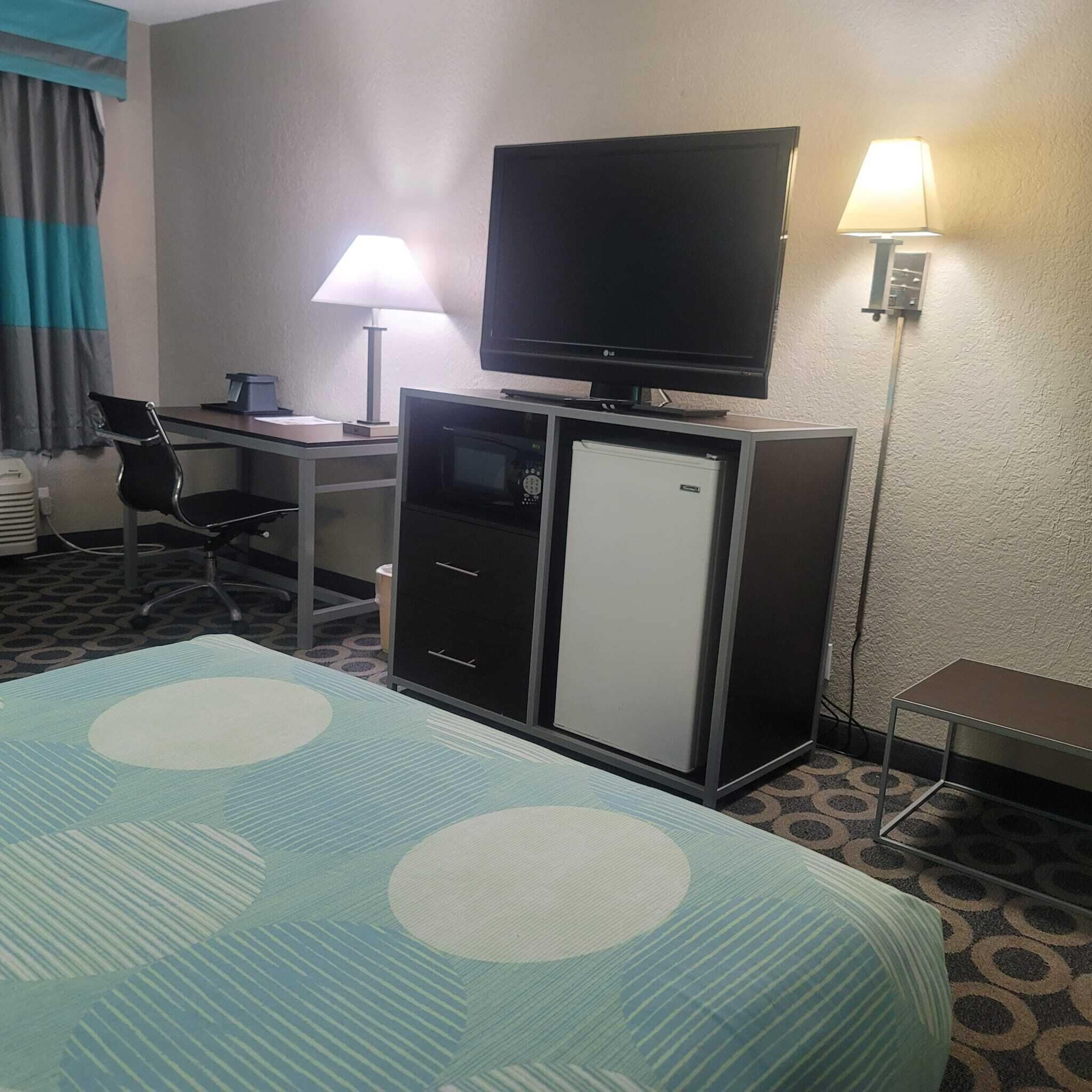 Habitación de hotel con cama, escritorio con computadora, televisión, refrigerador y dos lámparas.