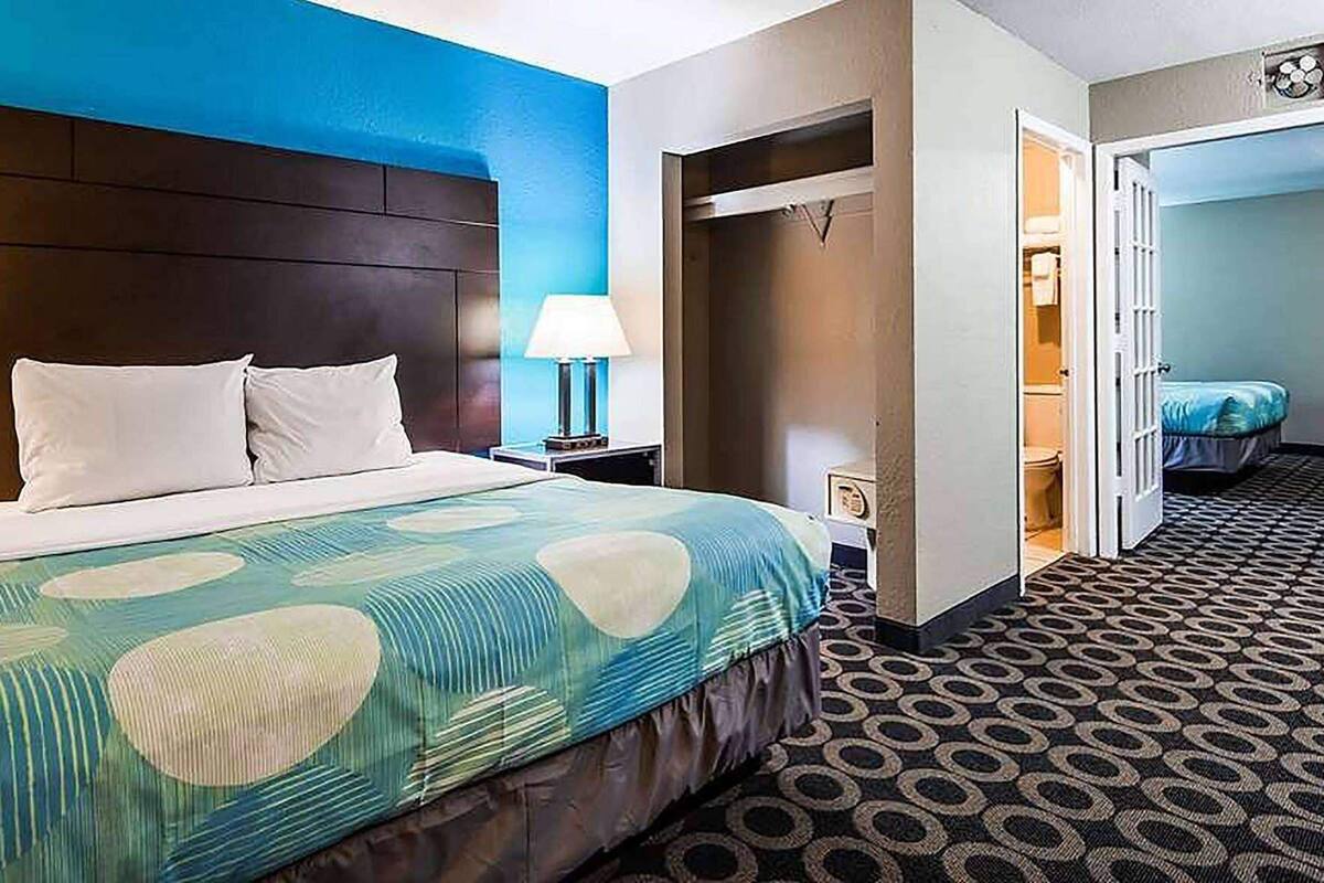 A hotel room with a bed, a nightstand, a lamp, a bathroom door, and a patterned carpet.