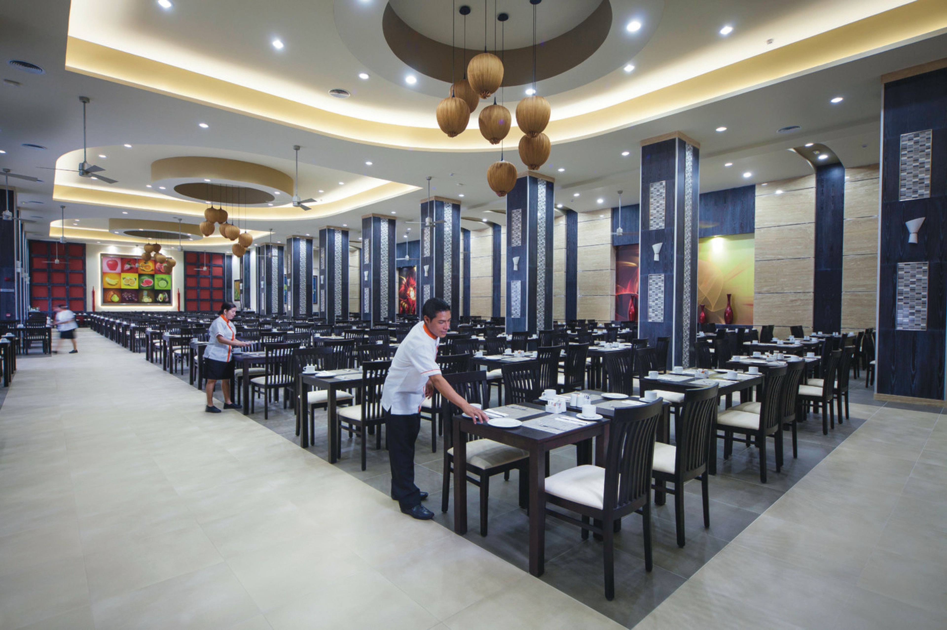 A spacious restaurant with multiple dining tables and chairs, a ceiling fan, and decorative hanging lights.
