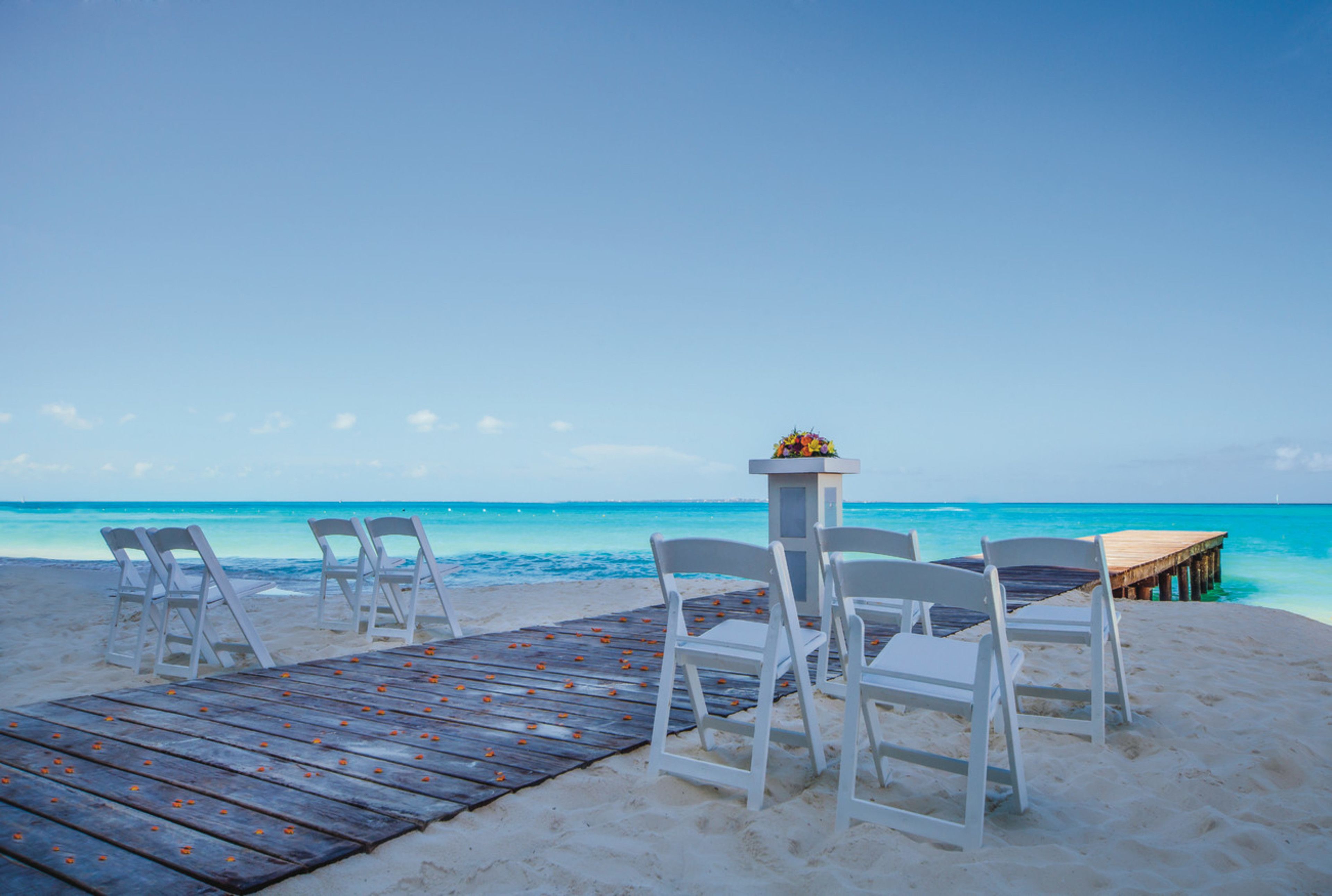 Een huwelijksceremonie op het strand met witte stoelen en een houten terras dat leidt naar een klein altaar met bloemen.