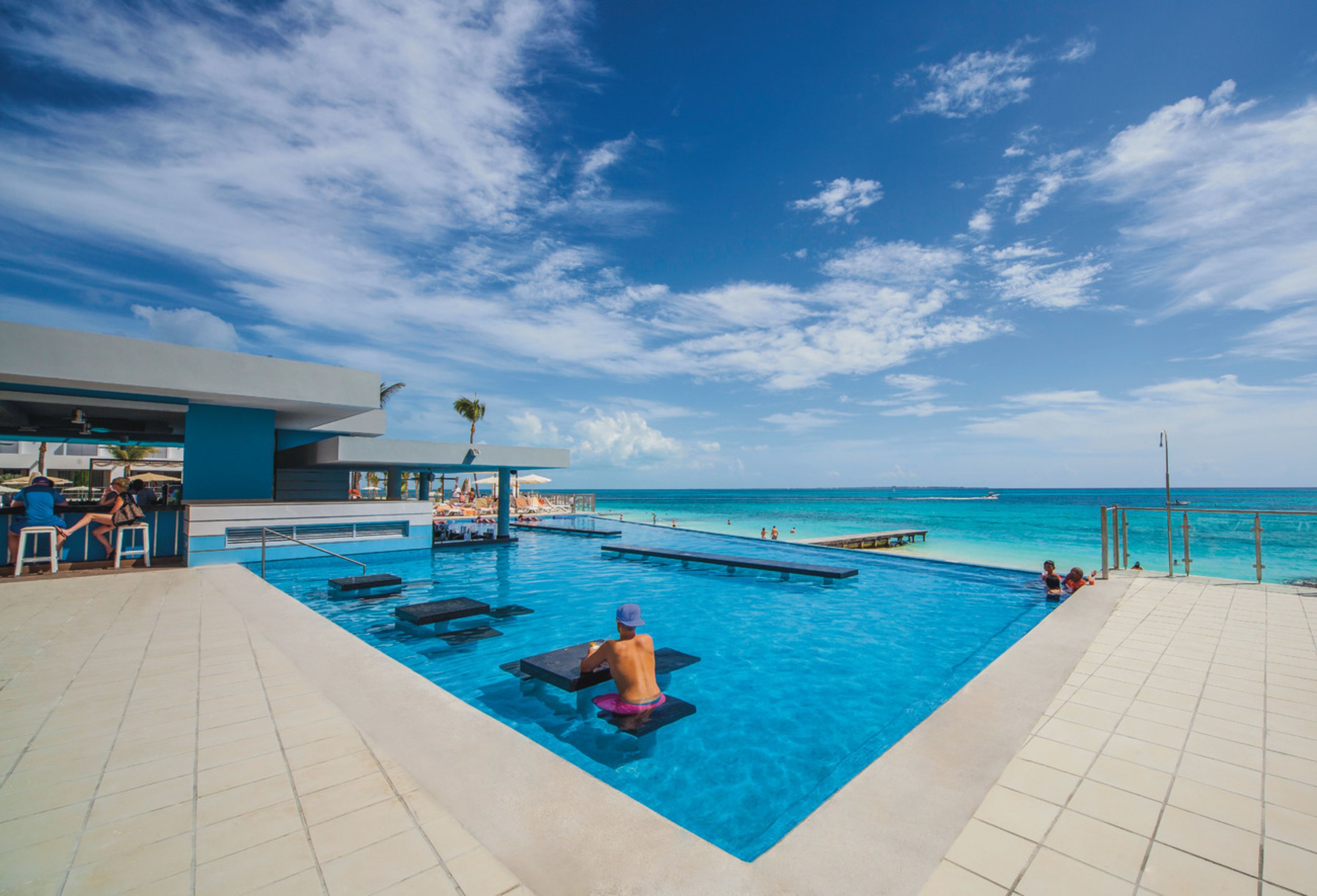 Área da piscina com vista para o oceano, cadeiras de sol e um bar.