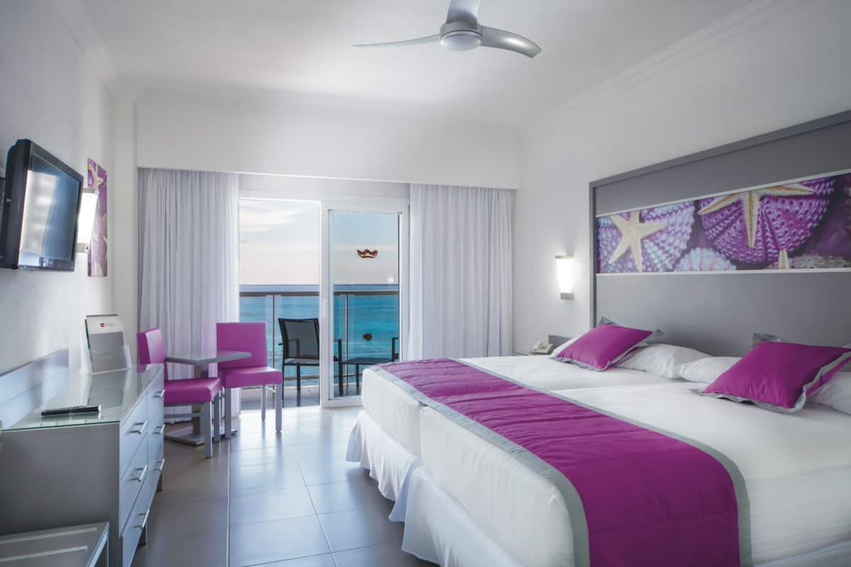 A hotel room with a bed, a desk, a chair, a TV, and a view of the sea.