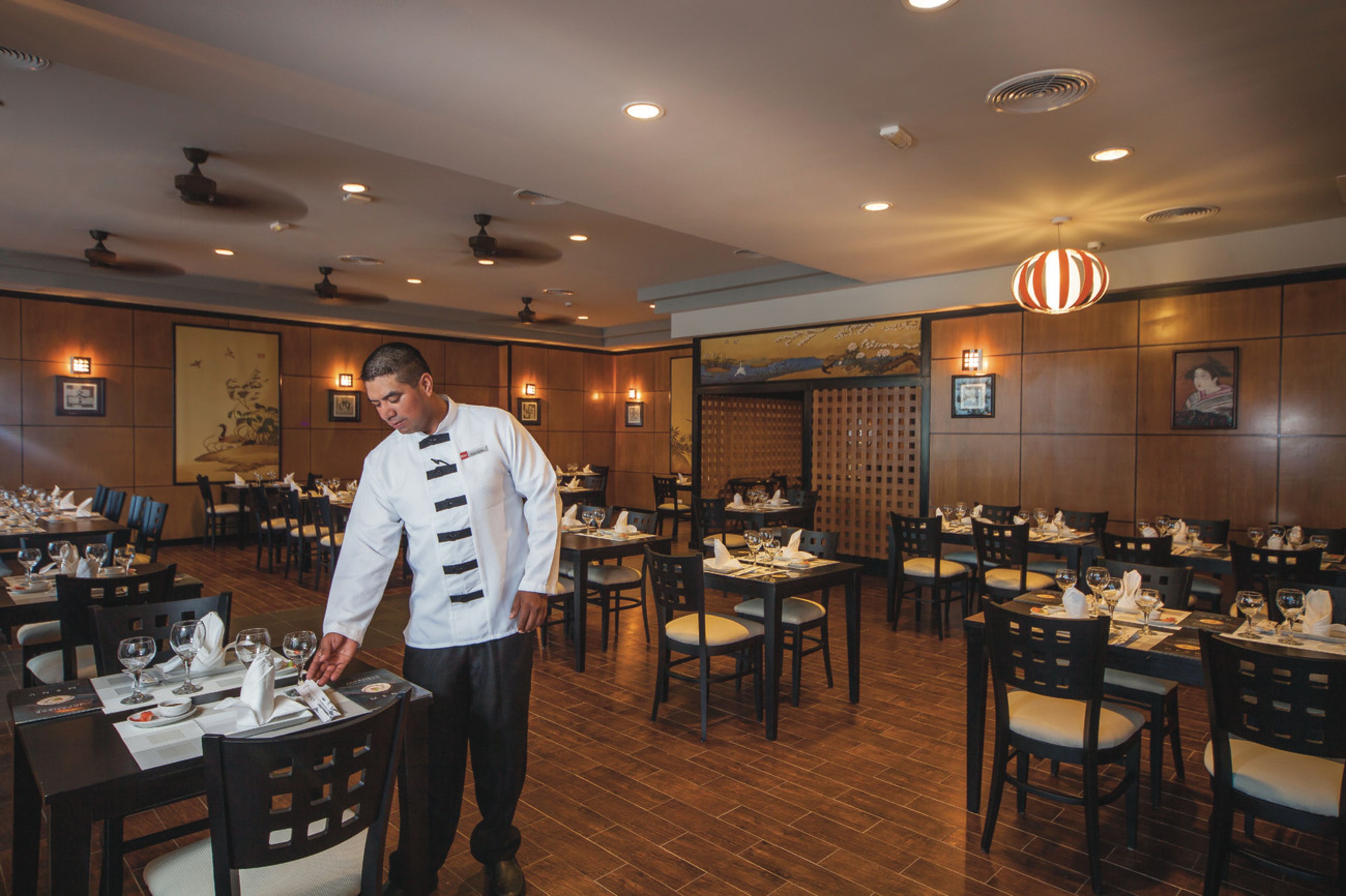A restaurant with wooden flooring, multiple tables set with white tablecloths and chairs, and a waiter in uniform.