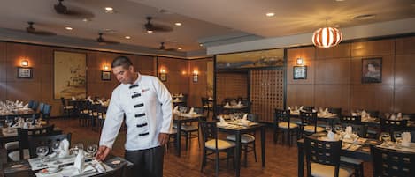 A restaurant with wooden flooring, multiple tables set with white tablecloths and chairs, and a waiter in uniform.