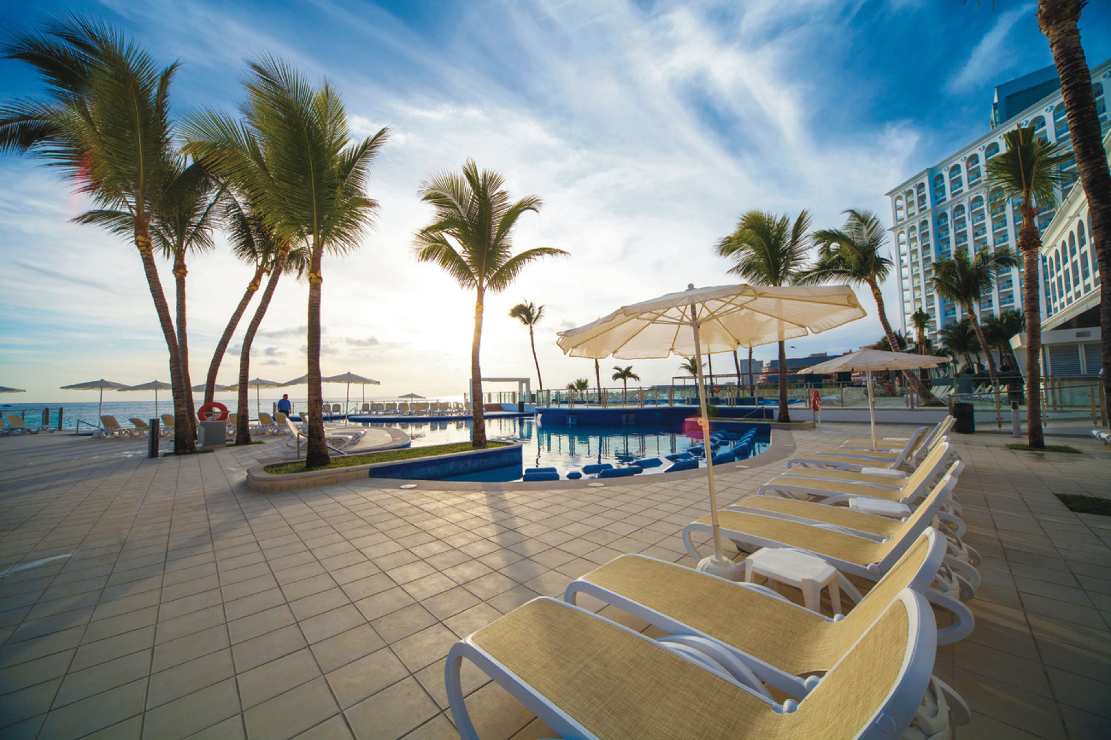 A poolside area with lounge chairs, umbrellas, and palm trees.