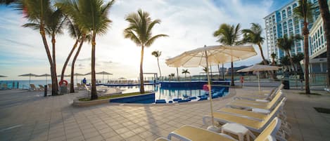 A poolside area with lounge chairs, umbrellas, and palm trees.