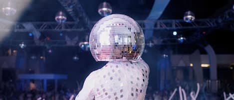 A person in a reflective, sequined outfit with a disco ball on their head, standing in front of a crowd at a concert or event.