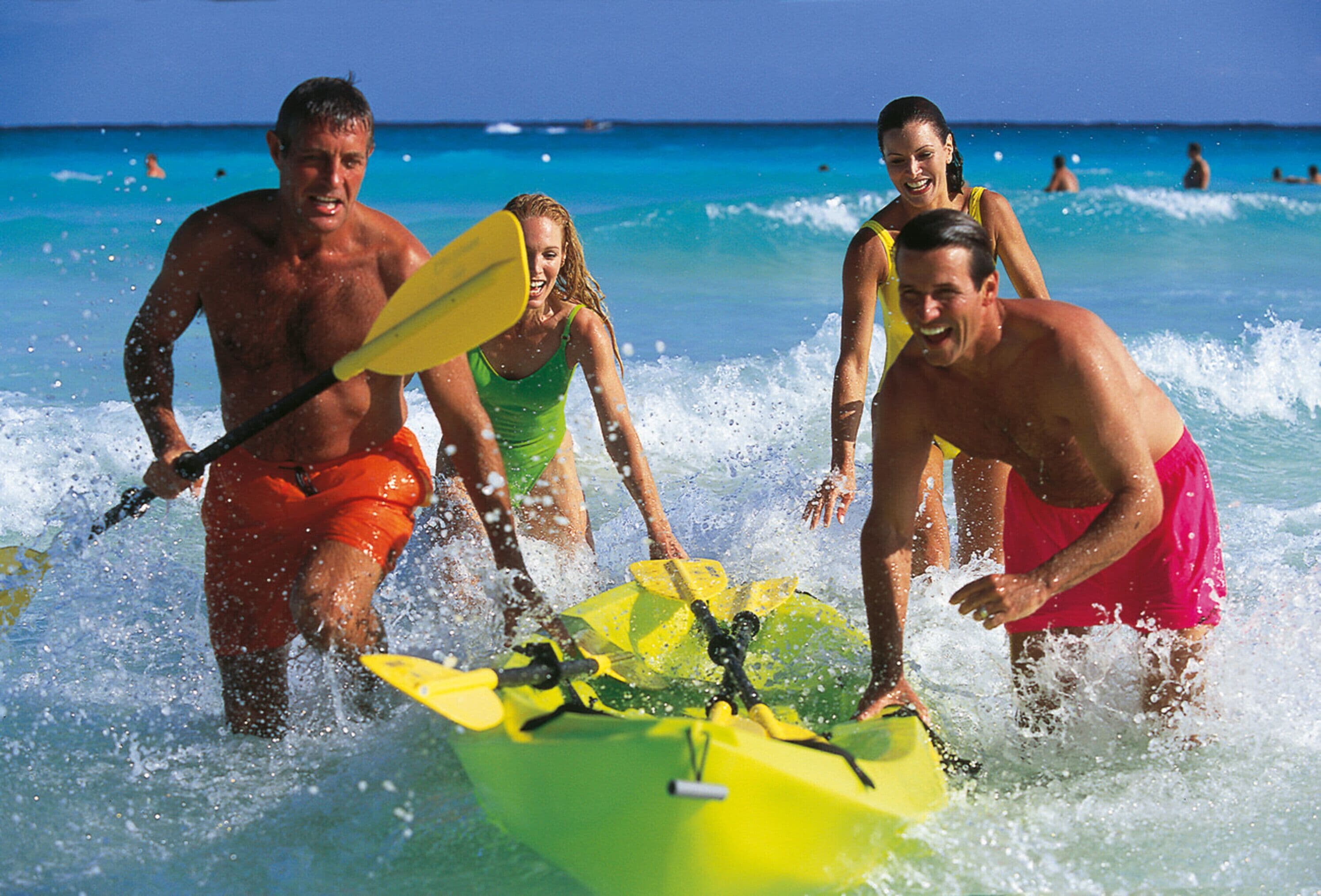 A group of people kayaking on a beach with clear blue water and a few other people in the background.