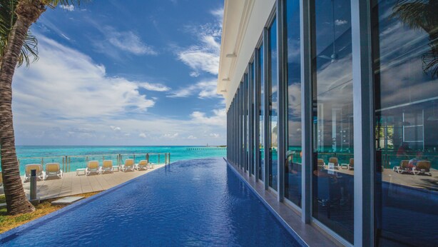 A poolside area with a view of the ocean, lounge chairs, and a modern building facade. - Hotel Riu Cancun -  Adults Only - All Inclusive (Cancun)
