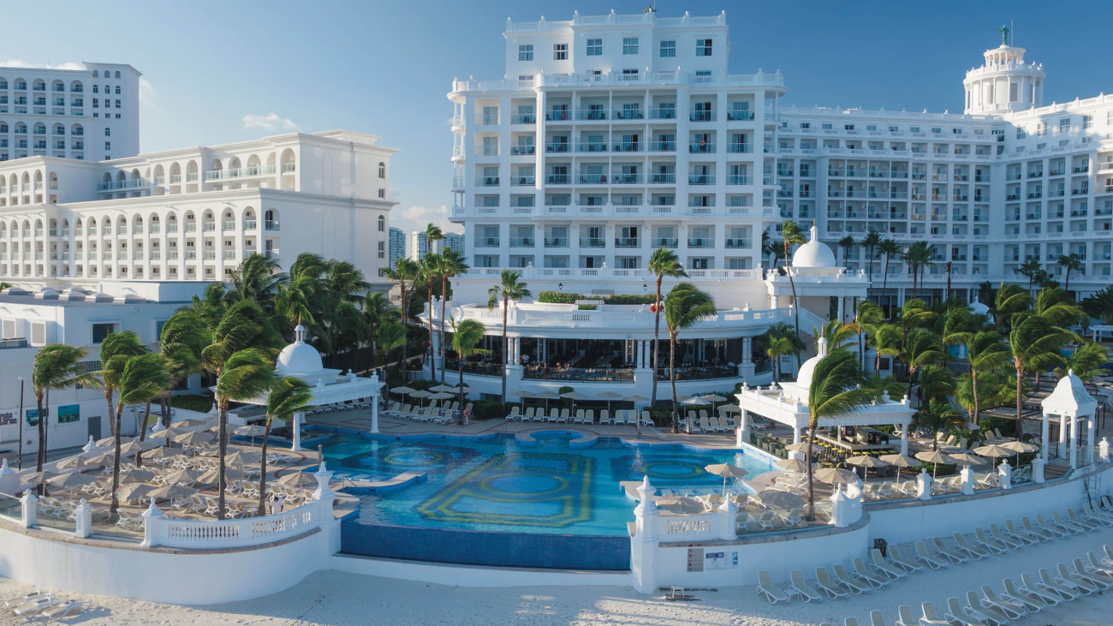 A large hotel with multiple floors, a central swimming pool, and surrounding palm trees.