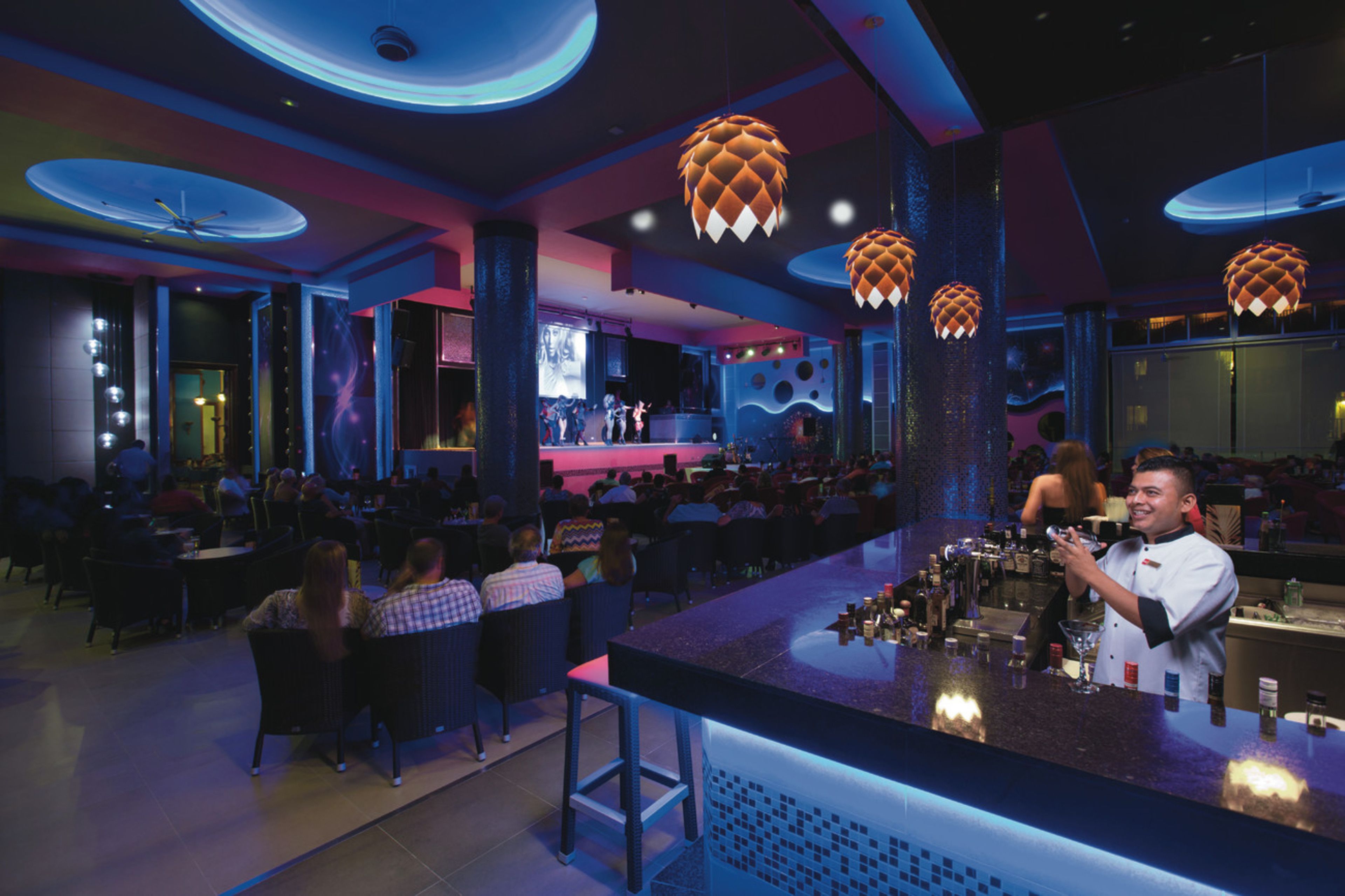 Zona de bar con un cantinero, mesas redondas y luces decorativas colgantes.