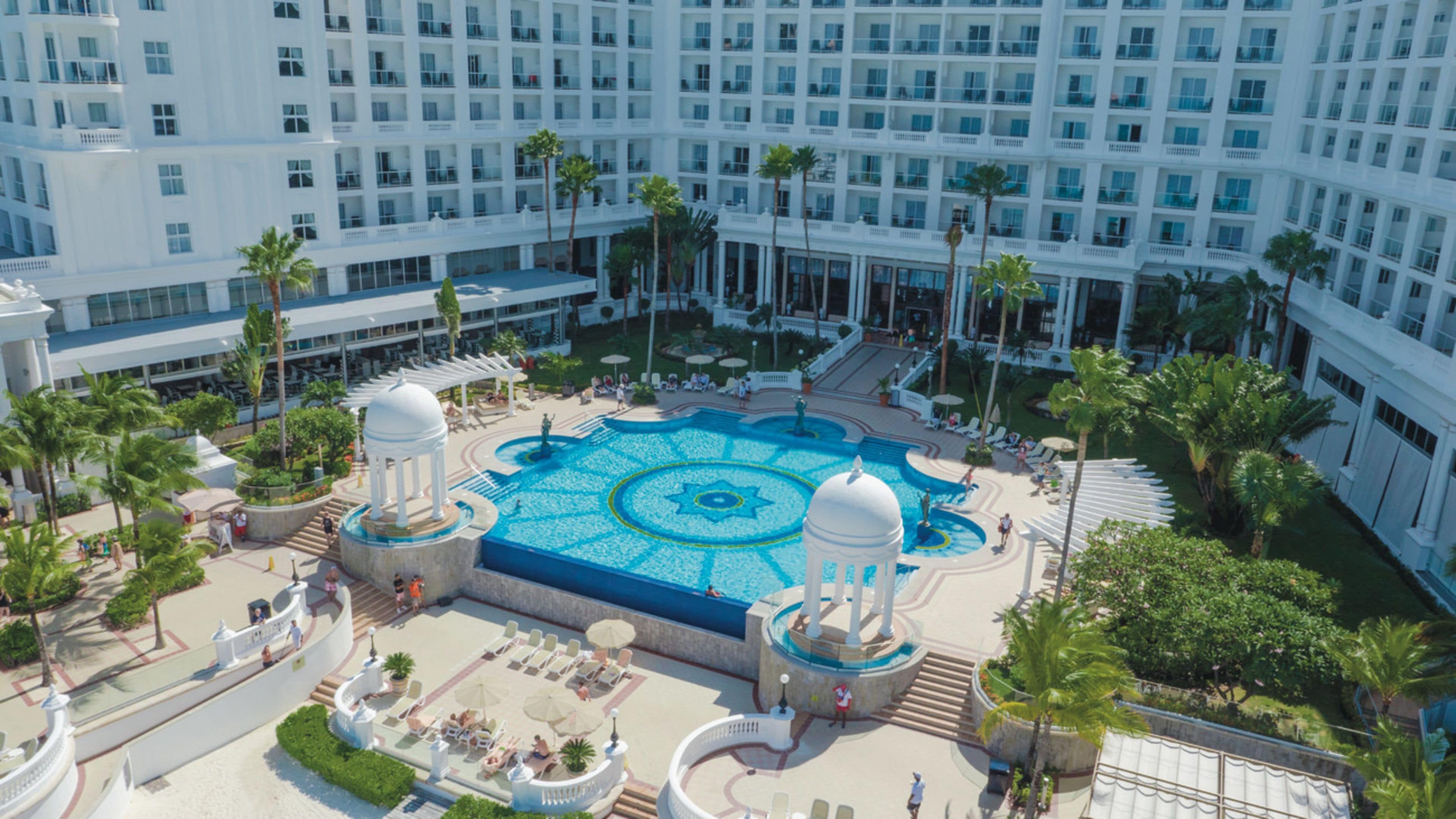 A hotel courtyard with a central pool, palm trees, and a large building in the background.