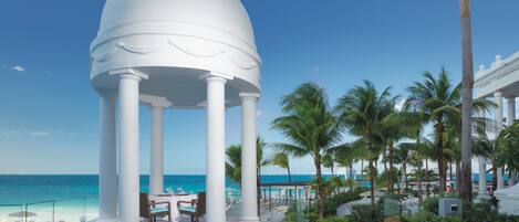 A white gazebo with columns, overlooking a beach with palm trees and a clear blue sky.