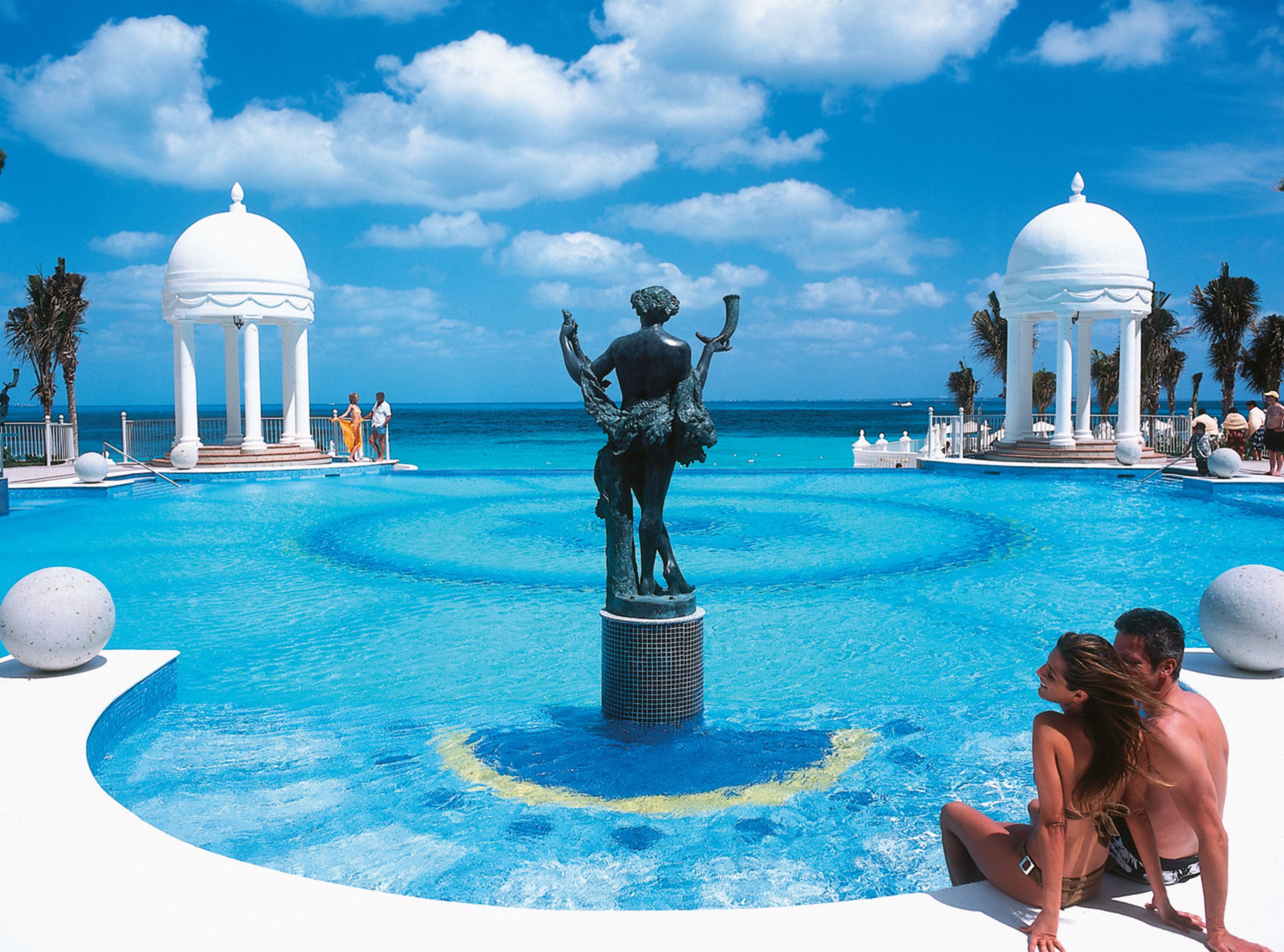 Una piscina con una estatua central, rodeada de estructuras blancas con cúpulas, palmeras y un cielo azul despejado.