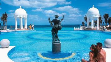 Una piscina con una estatua central, rodeada de estructuras blancas con cúpulas, palmeras y un cielo azul despejado.