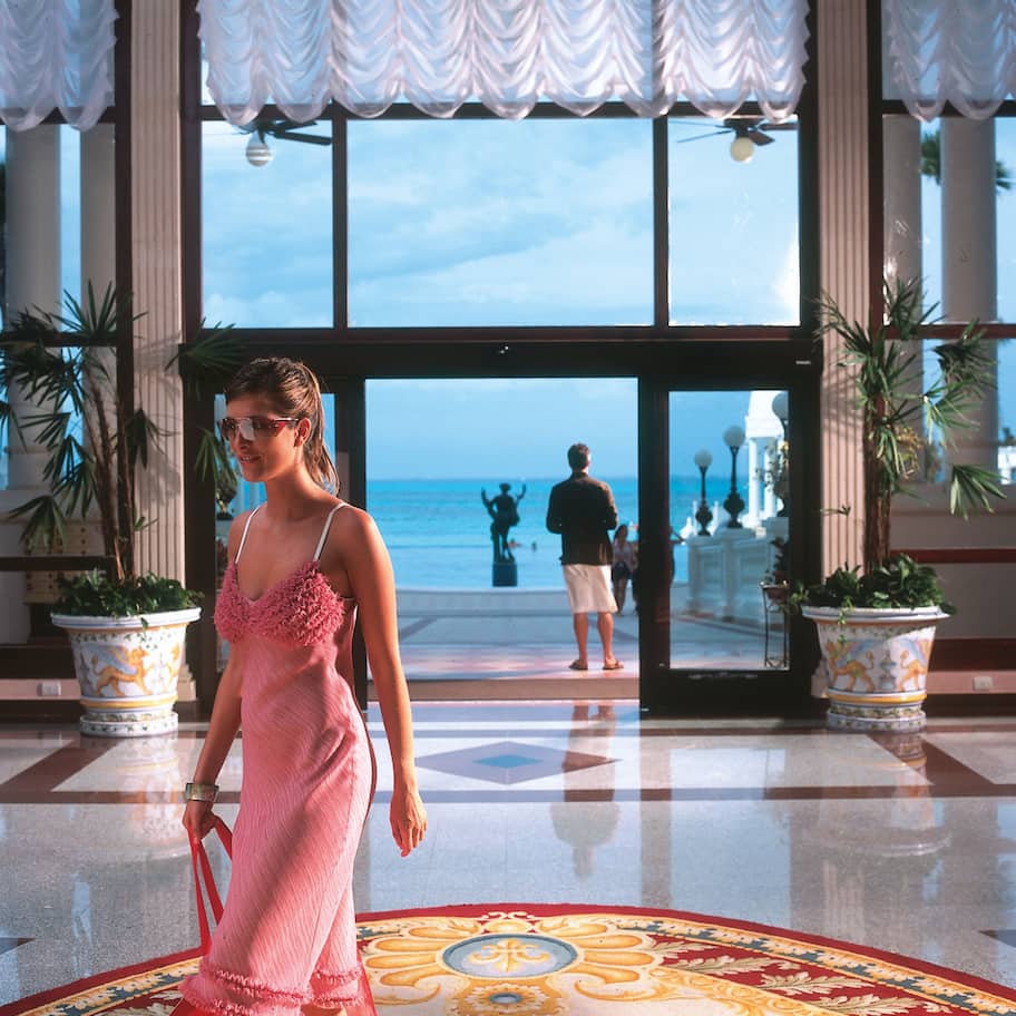 A woman in a pink dress walking through a lobby with large windows, potted plants, and a view of the sea.