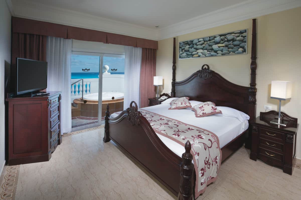 A hotel room with a large bed, a television, a nightstand, and a view of the beach through the sliding glass door.