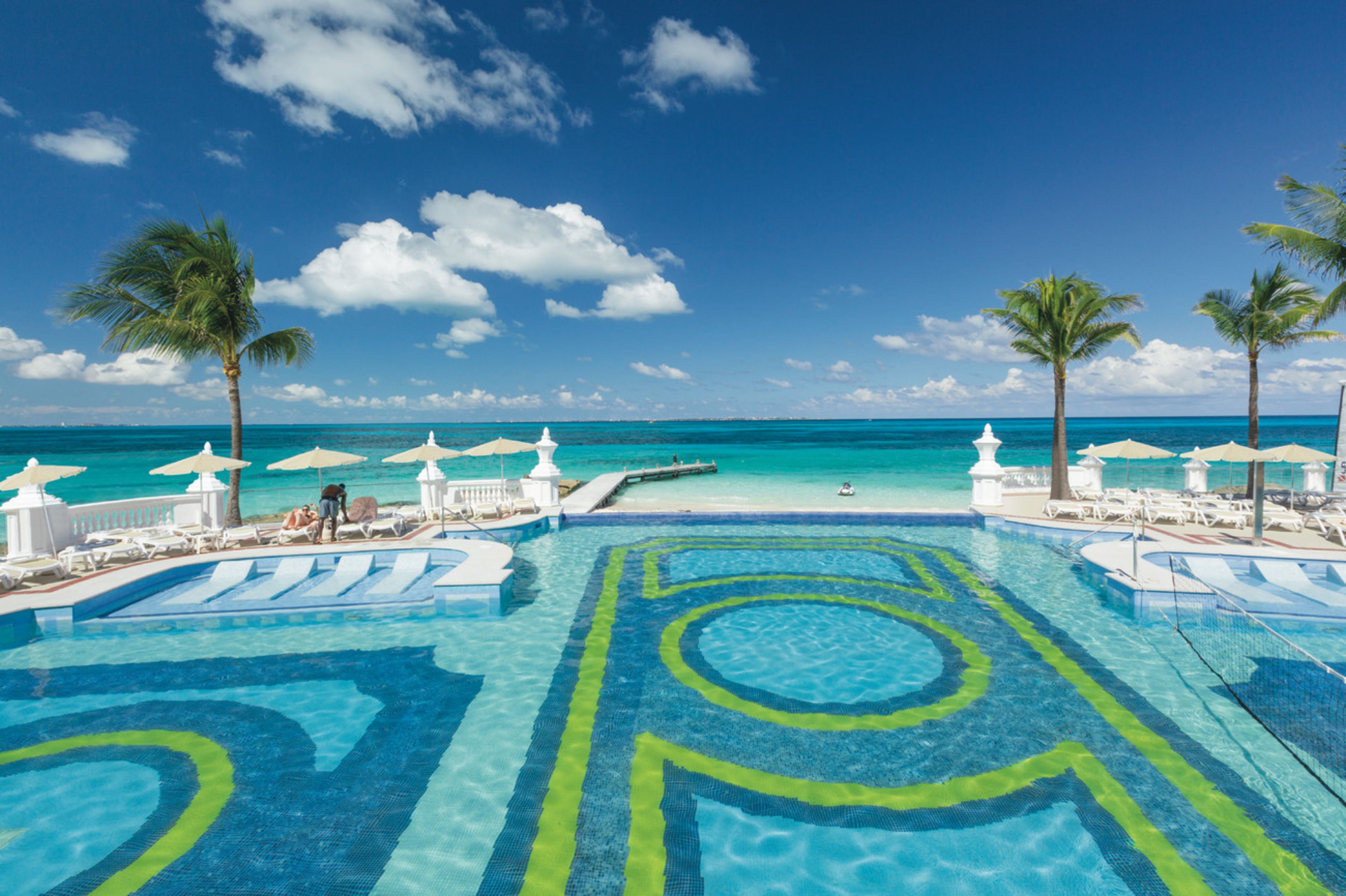 Una piscina con diseño tropical, rodeada de sillas de descanso y sombrillas, con vista al océano y palmeras.