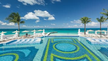 Una piscina con diseño tropical, rodeada de sillas de descanso y sombrillas, con vista al océano y palmeras.
