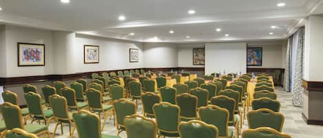 A conference room with rows of green chairs and tables, white walls, and framed artwork.