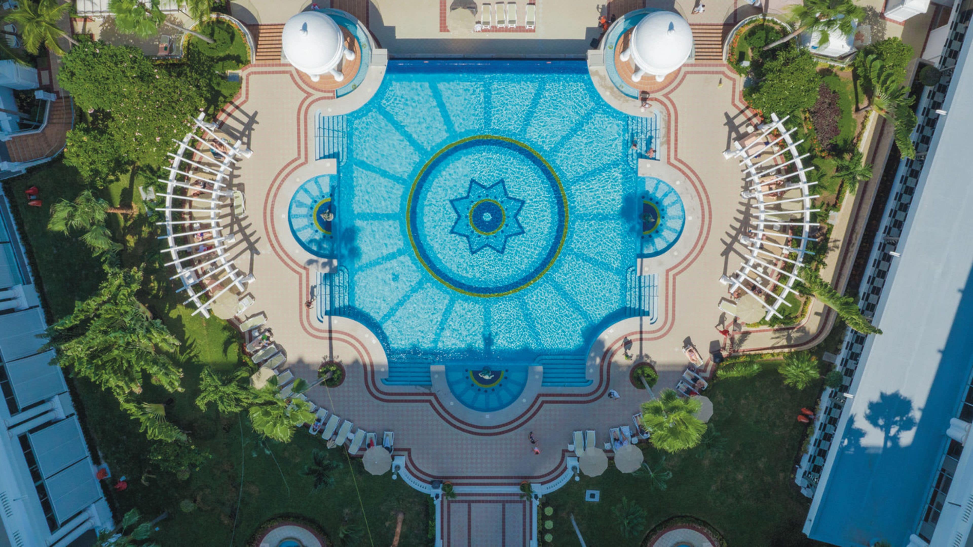Vista aérea de una piscina cuadrada con una fuente central, rodeada de sillas de descanso y palmeras.