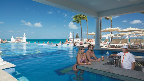 A poolside bar area with a view of the ocean, lounge chairs, and palm trees. - Hotel Riu Palace Las Americas - Adults Only- All Inclusive (Cancun)