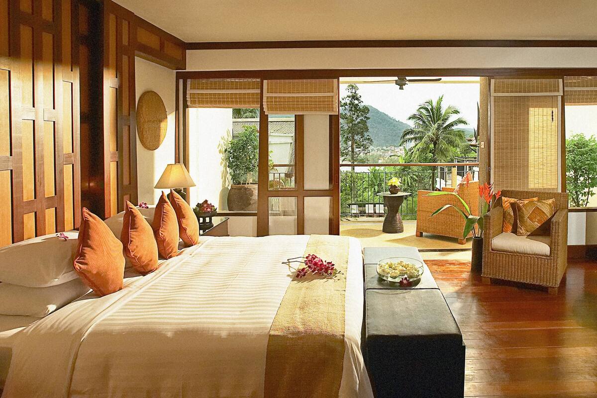 Habitación de hotel con cama, televisión, balcón con vistas a las montañas y zona de comedor.