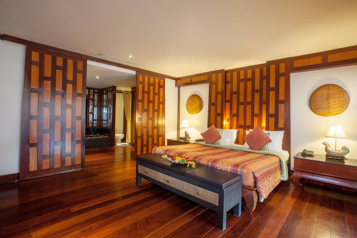 Habitación de hotel con piso de madera, cama con cubrecama estampado, dos mesitas de noche con lámparas, un banco con un arreglo floral y un panel decorativo en la pared.