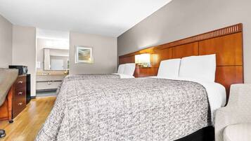 A hotel room with two beds, a wooden headboard, a bedside table with a lamp, a dresser, and a mirror.