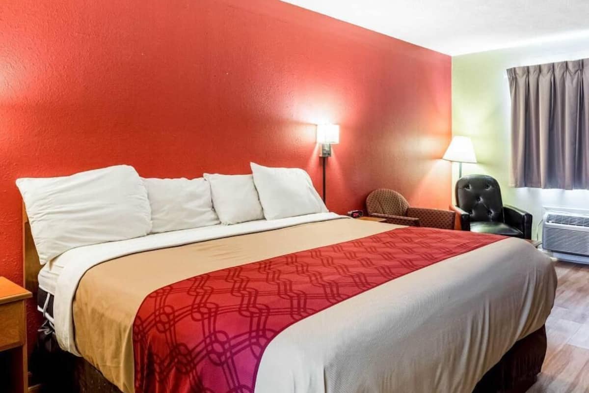 A hotel room with a bed, a nightstand, a chair, a lamp, and an air conditioning unit.