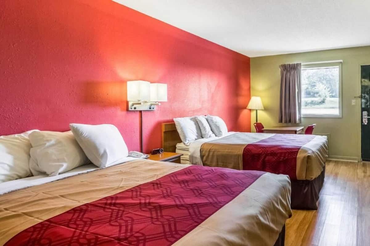 A hotel room with two beds, a red wall, a window with curtains, and a bedside table with a lamp.