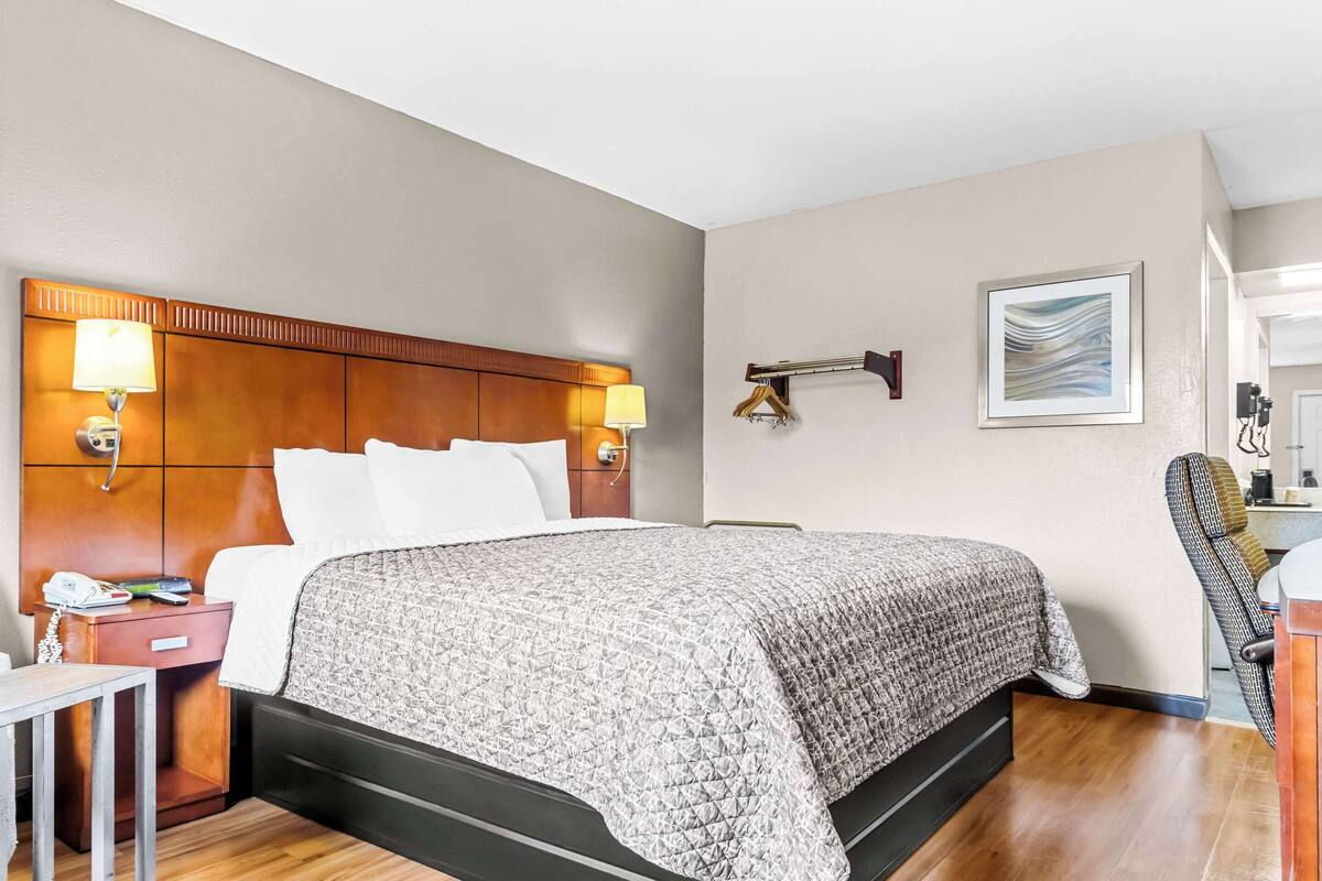 A hotel room with a large bed, bedside lamps, a wooden headboard, a small table with a phone, and a chair.
