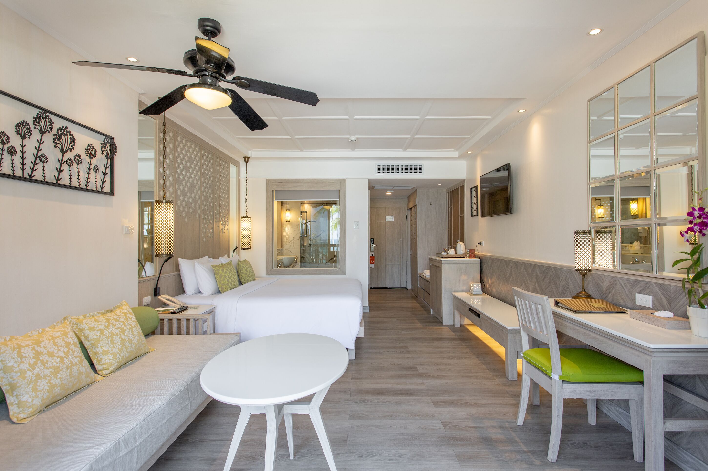 A hotel room with a bed, a desk, a chair, a table, a ceiling fan, a large mirror, and a decorative wall piece.
