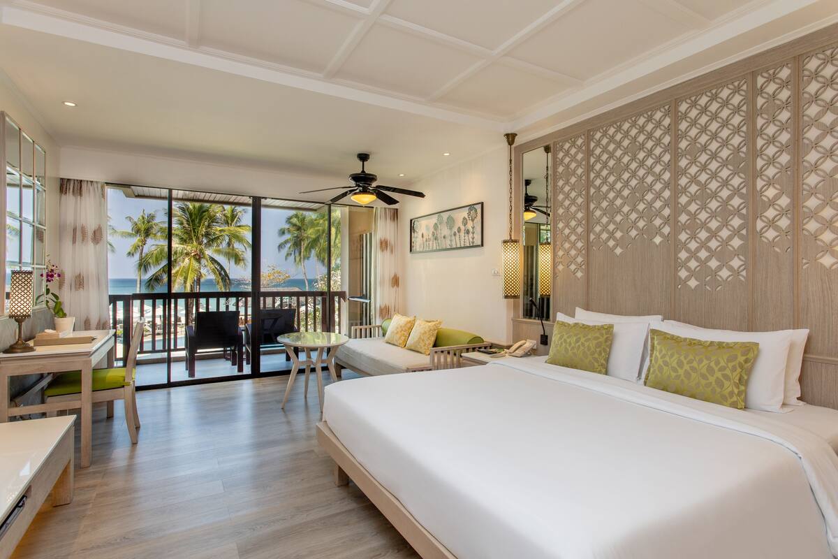 Ein Hotelzimmer mit einem großen Bett, einem Balkon mit Blick auf Palmen, einem Essbereich und einem Deckenventilator.