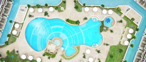 Vista aérea de un complejo turístico con una gran piscina central, varias piscinas más pequeñas y numerosas tumbonas y sombrillas.