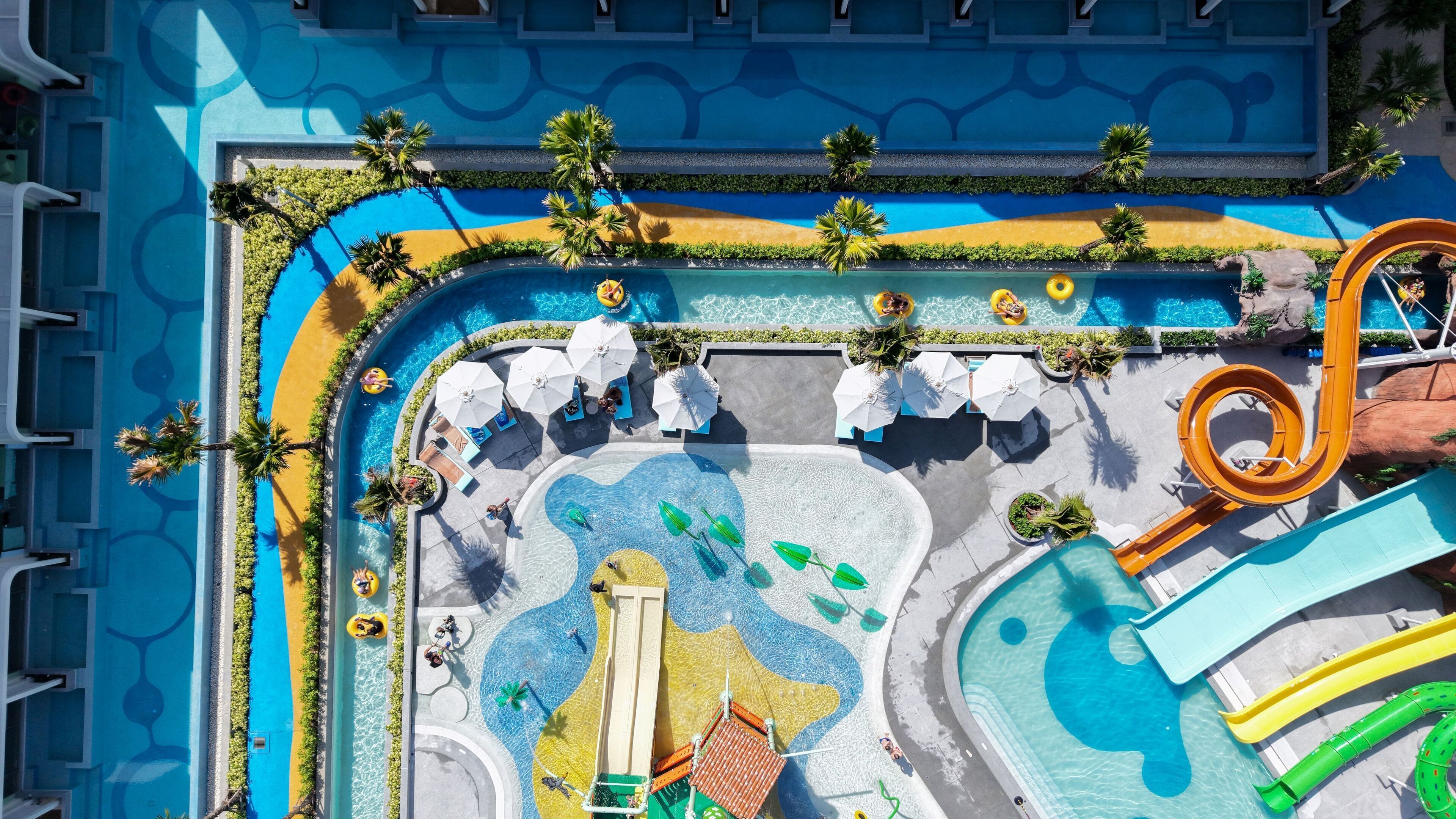 An aerial view of a resort pool area with multiple water slides, sun loungers, and palm trees.