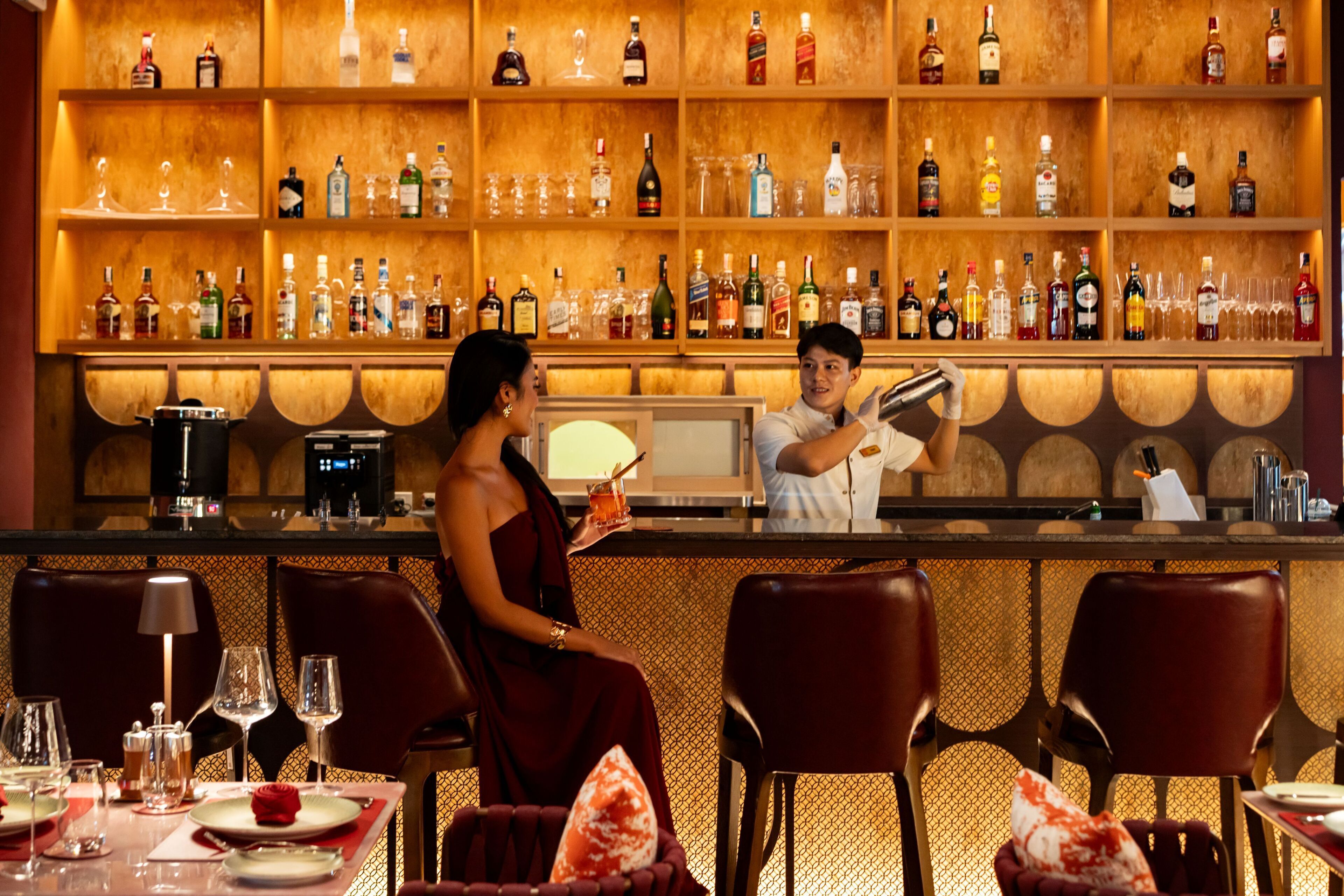 A bar area with shelves of liquor bottles and glasses, a bartender preparing a drink, and a seated woman holding a drink.