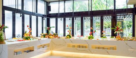 A spacious, well-lit room with large windows, a marble counter with various fruits and flowers, and a clean, modern design.