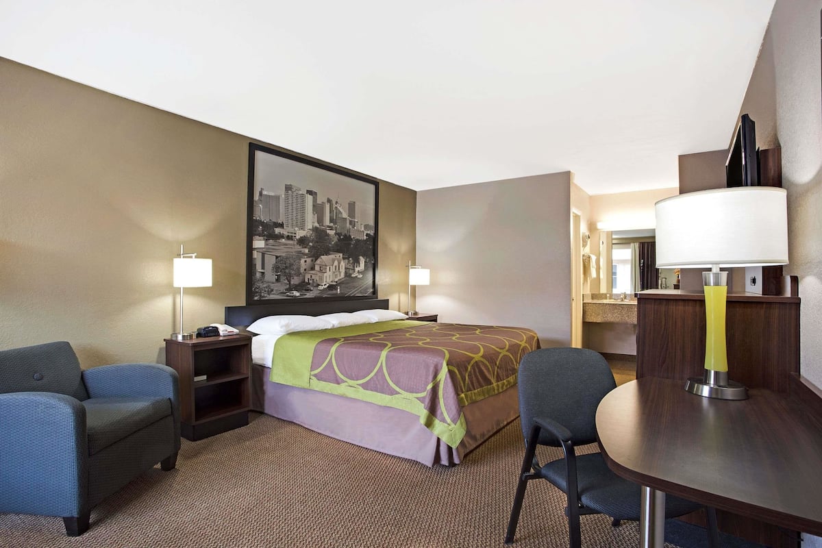 A hotel room with a bed, two chairs, a desk, and a large cityscape painting on the wall.