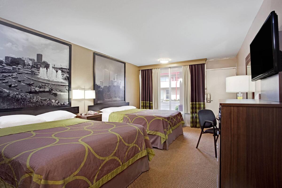 A hotel room with two beds, a television, a desk, and a large window with a cityscape view.