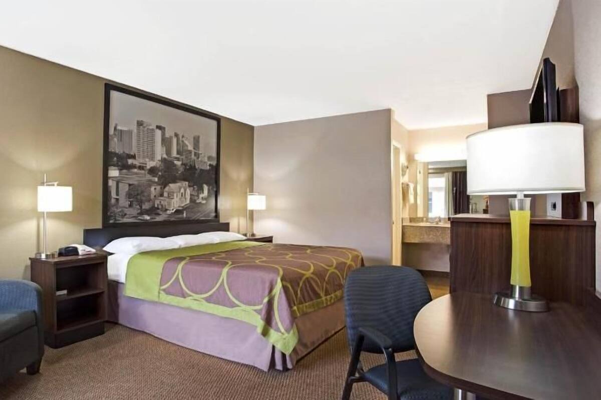 A hotel room with a bed, a desk, a chair, a sofa, two bedside tables with lamps, and a large cityscape painting on the wall.